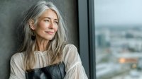 Confident mature Asian woman with long gray hair in elegant beige blouse looking thoughtfully out office window with city view background.