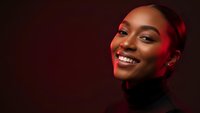 Young African American woman with radiant smile and glowing skin in warm red lighting against dark background for beauty and lifestyle campaigns.