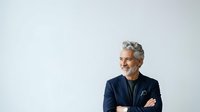 Mature man with gray hair and facial hair wearing navy blazer with pocket square, arms crossed, looking away thoughtfully against white background for corporate and business use.