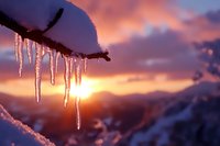 Dramatic icicles hanging from frozen branch against vibrant sunset sky over snow-covered mountain landscape during winter season creating magical atmosphere.
