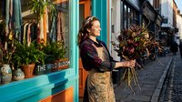 Young woman with brown hair and fair complexion standing outside a turquoise storefront holding a dried flower bouquet, wearing brown jacket and patterned skirt for florist, retail, small business, lifestyle.