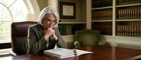 Senior Caucasian female lawyer in contemplative pose at mahogany desk in traditional law office with legal books and documents for professional consultation.