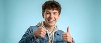 Happy young Caucasian man with curly hair giving double thumbs up gesture in denim jacket against blue background for positive marketing campaigns.