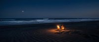 Romantic couple sitting by campfire on beach at twilight with ocean waves and starry sky creating intimate evening atmosphere for vacation getaway.
