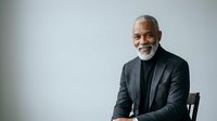 Mature African American male professional with gray beard wearing dark blazer and black turtleneck, seated against neutral background for corporate portraits and business headshots.