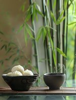 Asian dumplings in ceramic bowl with tea cup on bamboo mat, green bamboo plants in background creating zen atmosphere.