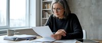 Focused mature businesswoman with gray hair and glasses reviewing documents at office desk near window with natural lighting for professional workflow.