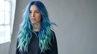 Young woman with vibrant blue and teal ombre hair color standing in modern interior with natural window light for beauty and fashion concepts.
