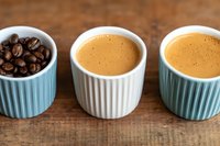 Three ceramic ramekins on wooden surface, one filled with roasted coffee beans and two with freshly brewed espresso shots topped with golden crema.