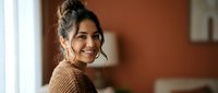 Young Hispanic woman with messy bun hairstyle wearing brown knit sweater smiling warmly at camera in cozy home interior with natural lighting.