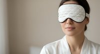Young Asian woman wearing white quilted sleep mask and bathrobe for relaxation and rest in bedroom setting.