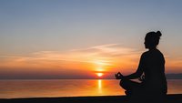 Woman practicing yoga meditation in lotus pose silhouette against golden sunset over calm water creating peaceful mindfulness moment for wellness and relaxation.