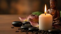 Zen spa composition with Buddha statue, burning candle, hot stones and lotus flower creating peaceful meditation atmosphere for wellness and relaxation.