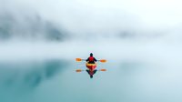 Lone kayaker in red jacket paddling on misty lake with mirror reflection, creating minimalist composition in cool blue tones. Peaceful outdoor scene.