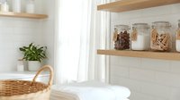 Bright, minimalist bathroom interior with white subway tile walls, wooden floating shelves holding glass storage jars with dried goods, potted green plant, woven basket, and natural window light for home decor and organization inspiration.