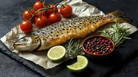 Fresh raw trout with crispy scales on parchment paper, garnished with cherry tomatoes on vine, lime slices, rosemary sprigs and mixed peppercorns in ceramic bowl.