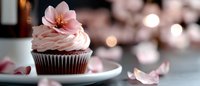Chocolate cupcake with pink buttercream frosting topped with delicate flower decoration on white plate. Soft bokeh lights create romantic atmosphere for bakery menu.