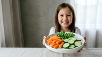 Happy young girl holding plate of fresh vegetables including carrots and cucumbers promoting healthy eating habits for children nutrition education.