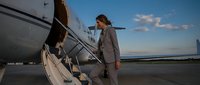 Confident young Caucasian businesswoman in gray suit boarding private jet on airport tarmac at dusk, conveying luxury travel and success.