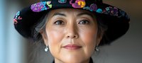 Mature Asian woman wearing elegant black hat decorated with colorful floral embroidery, pearl earrings, and gentle smile in natural lighting.