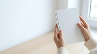 Woman holding blank white tablet device on wooden table with bright window background, mockup for app design and digital marketing.