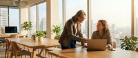 Two professional businesswomen collaborating on laptop in modern office with city view. Corporate teamwork and mentorship in bright workspace.