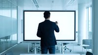 Professional businessman in dark suit presenting to blank white screen in modern glass conference room with contemporary office furniture and natural lighting.