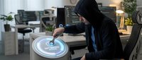 Hooded hacker in dark clothing accessing futuristic holographic interface in modern office environment with multiple computer screens and technology.
