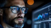 Caucasian businessman with beard and glasses analyzing financial market data on computer screen in dark office, illuminated by blue monitor glow.