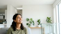 Young woman with dark hair and olive complexion wearing olive-green shirt, sitting indoors in bright modern home with potted plants on shelves, natural light from windows, contemplative expression looking upward.