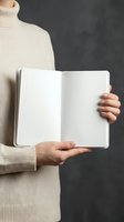 Woman in beige turtleneck sweater holding open blank notebook with empty white pages against dark background for mockup and design projects.