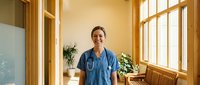 Middle-aged woman with light skin tone wearing blue medical scrubs in bright, modern clinic or hospital office with wooden frames and plants.