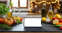 Kitchen counter with fresh vegetables, herbs, olive oil bottles and blank smartphone screen for cooking apps or recipe display in warm cozy home setting.