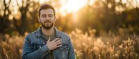 Caucasian male with beard wearing denim shirt standing in golden wheat field at sunset, hand on chest, expressing gratitude or sincerity in natural outdoor setting.