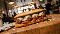 Gourmet meatball sandwich with melted cheese and tomato sauce on crusty bread, served on wooden cutting board in casual restaurant setting.