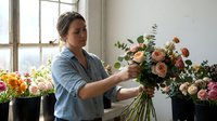 Woman florist arranging fresh cut flowers in vase by bright window, creating bouquet with roses and greenery for floral design work.