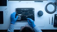 Technician in blue gloves repairing laptop circuit board on white workbench with tools and components, overhead view for tech support and electronics maintenance services.
