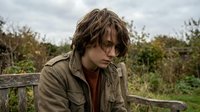 Sad teenage Caucasian boy with messy brown hair sitting on bench outdoors, looking down, wearing olive jacket, overcast sky, green nature background.