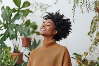 Young African American woman with natural curly hair smiling with eyes closed, wearing brown sweater, surrounded by green plants against white wall background.