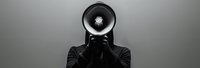 Person in black clothing holding megaphone in front of face against gray background, creative conceptual photography representing communication and protest.