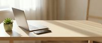 Laptop and tablet on wooden desk with small potted plant and window light, workspace setup for remote work and digital productivity.
