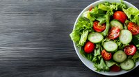 Fresh garden salad with sliced cucumbers, cherry tomatoes and green lettuce leaves served in white bowl on dark wooden background. Top view.