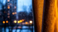 Raindrops on window glass with blurred city lights at dusk, warm orange curtain creating cozy atmosphere. Moody evening urban scene through wet window pane.