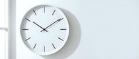 Modern minimalist white wall clock with clean lines showing 10:10 time on bright white wall near window for contemporary home office interior design.