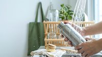 Hands holding reusable glass water bottle in sustainable eco-friendly kitchen with bamboo accessories and green shopping bag for zero waste lifestyle.