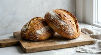 Two freshly baked artisan sourdough bread loaves with golden crusts on rustic wooden cutting board by window for bakery and homemade cooking concepts.