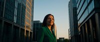 Confident businesswoman in green suit standing between modern glass skyscrapers at sunset, smiling with ambition and success.