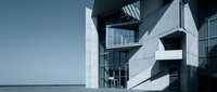 Contemporary waterfront building with angular white facade and geometric balconies overlooking calm sea under clear sky. Suitable for architecture, real estate, and modern design projects.