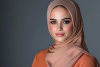 Young Middle Eastern woman wearing beige hijab and orange top smiling confidently against dark background, representing modern Muslim professional lifestyle.