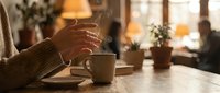 Woman hands holding warm coffee mug in cozy cafe atmosphere with soft lighting and plants creating peaceful morning moment for relaxation and comfort.